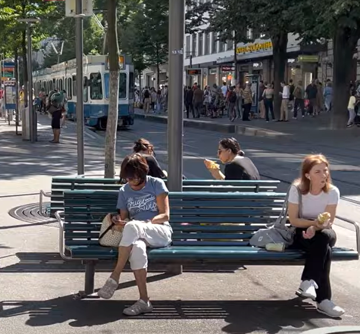 Two people sitting on a bench on a sunny day. In the background, a tram is visible. Many people standing on the sidewalk. 