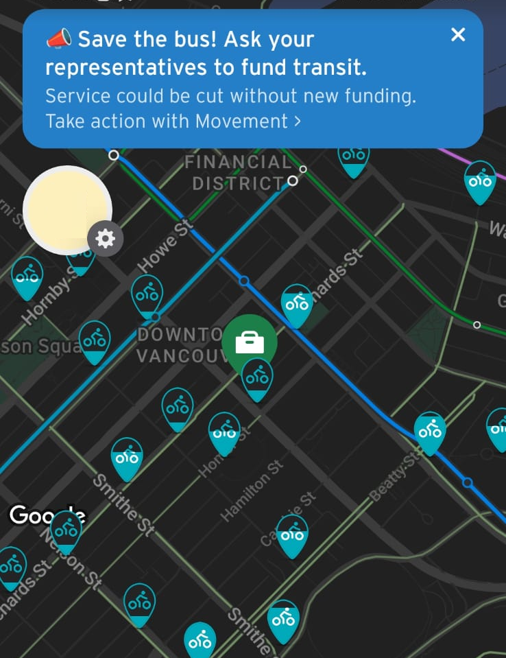 The image is a screenshot of a transit navigation app displaying a map of downtown Vancouver. The map shows blue lines representing transit routes and multiple icons indicating bike-sharing stations. A blue notification banner at the top reads: "🚨 Save the bus! Ask your representatives to fund transit. Service could be cut without new funding. Take action with Movement The background map is dark, with street names and landmarks labeled in white. A circular navigation icon is on the left side, and a green icon with a white briefcase symbol is present, possibly indicating a specific location or service. The image emphasizes the need for transit funding and encourages users to take action.