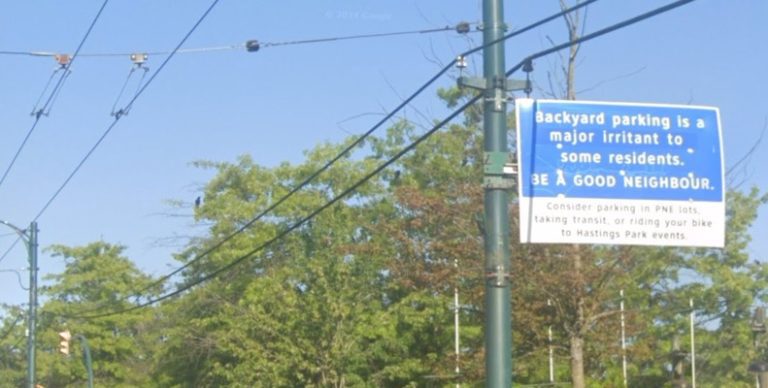 A screenshot from google maps of a blue and white sign that says "backyard parking is a major irritant to some residents. BE A GOOD NEIGHBOUR." on a blue background and a white highway font "Consider parking in PNE lots, taking transit or riding your bike to Hastings Park events." in a black highway font on a white background there are trees and trolley wires in the background