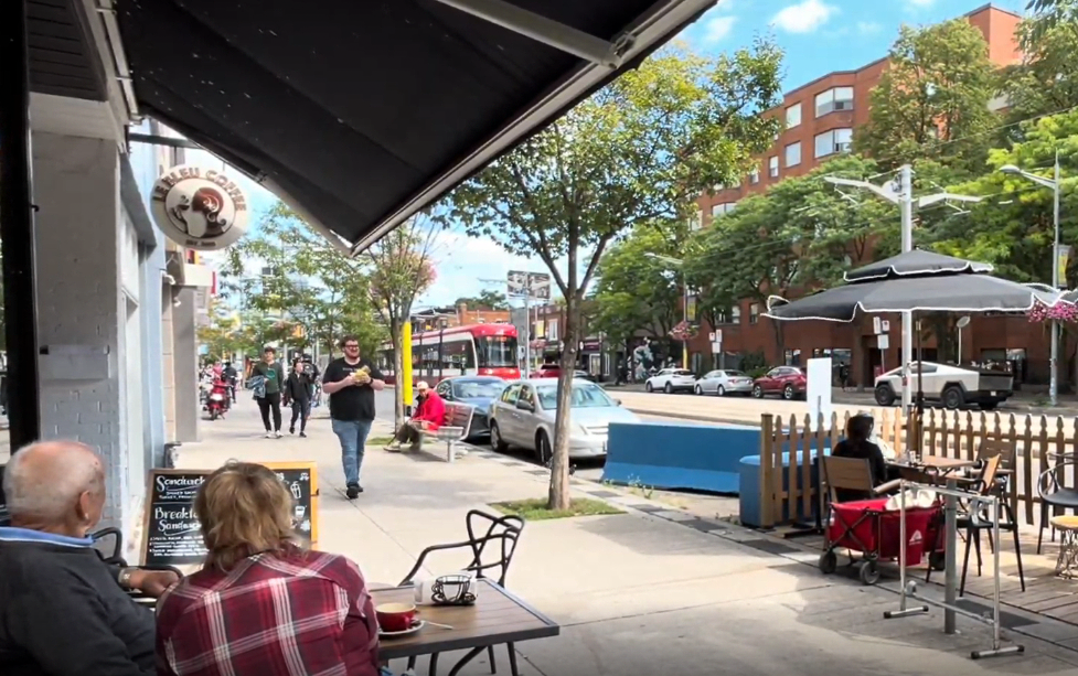 Toronto’s St. Clair West is a leafy main street, the photo is from a nice day with people sitting at a sidewalk table having coffee, along with many others walking down the sidewalk. A transit vehicle glides through dedicated lanes in the background, while parked cars share a lane with more patios in the curb lane.